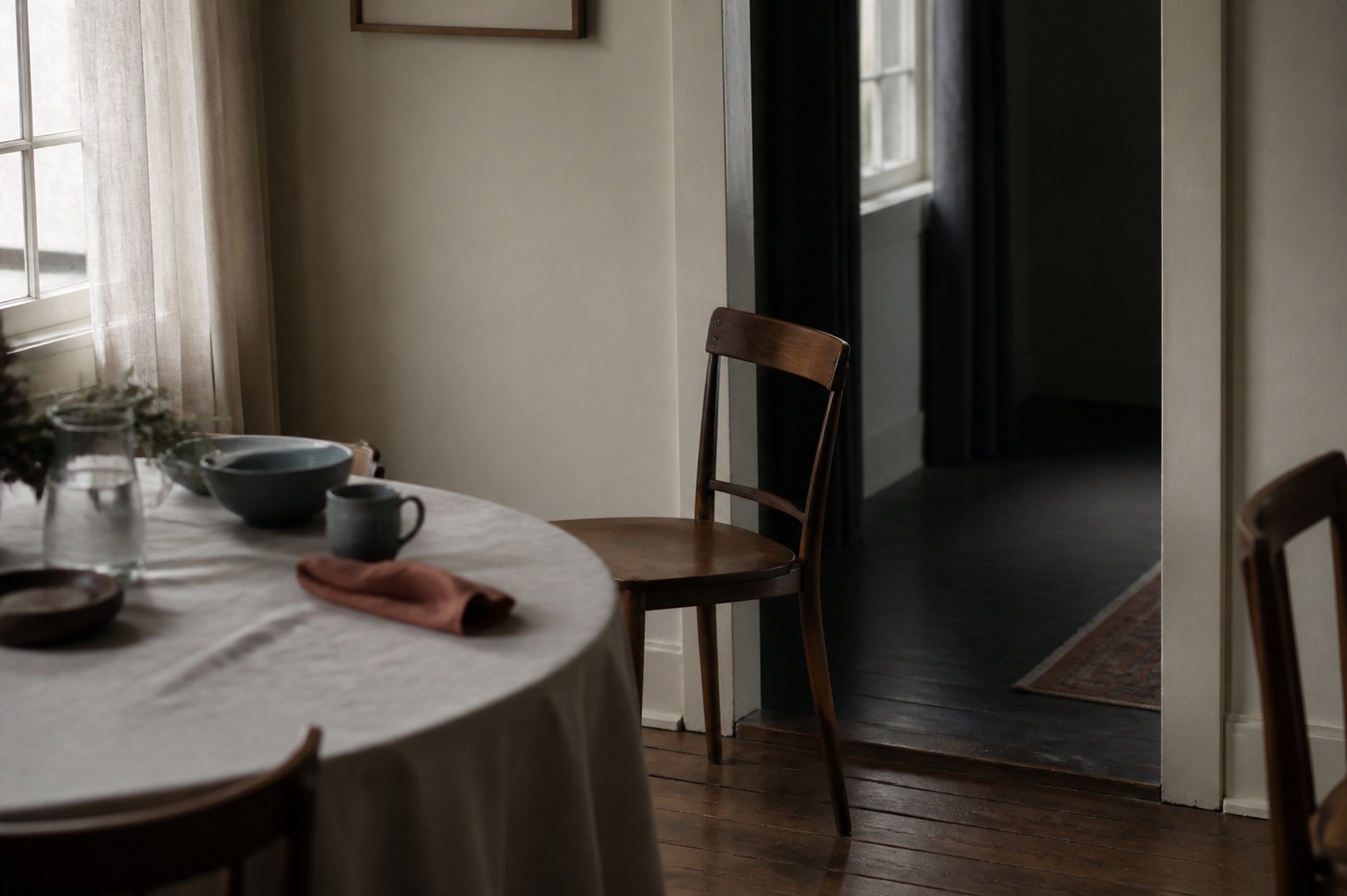 An empty chair sits partly at a set table and partly in the passage to a darker adjoining room.